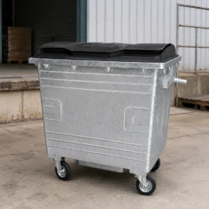 1100L Galvanized Steel Waste Container for Industrial Sites | KOVANTO 1100L galvanized steel waste container with black plastic lid and four caster wheels in an industrial loading area
